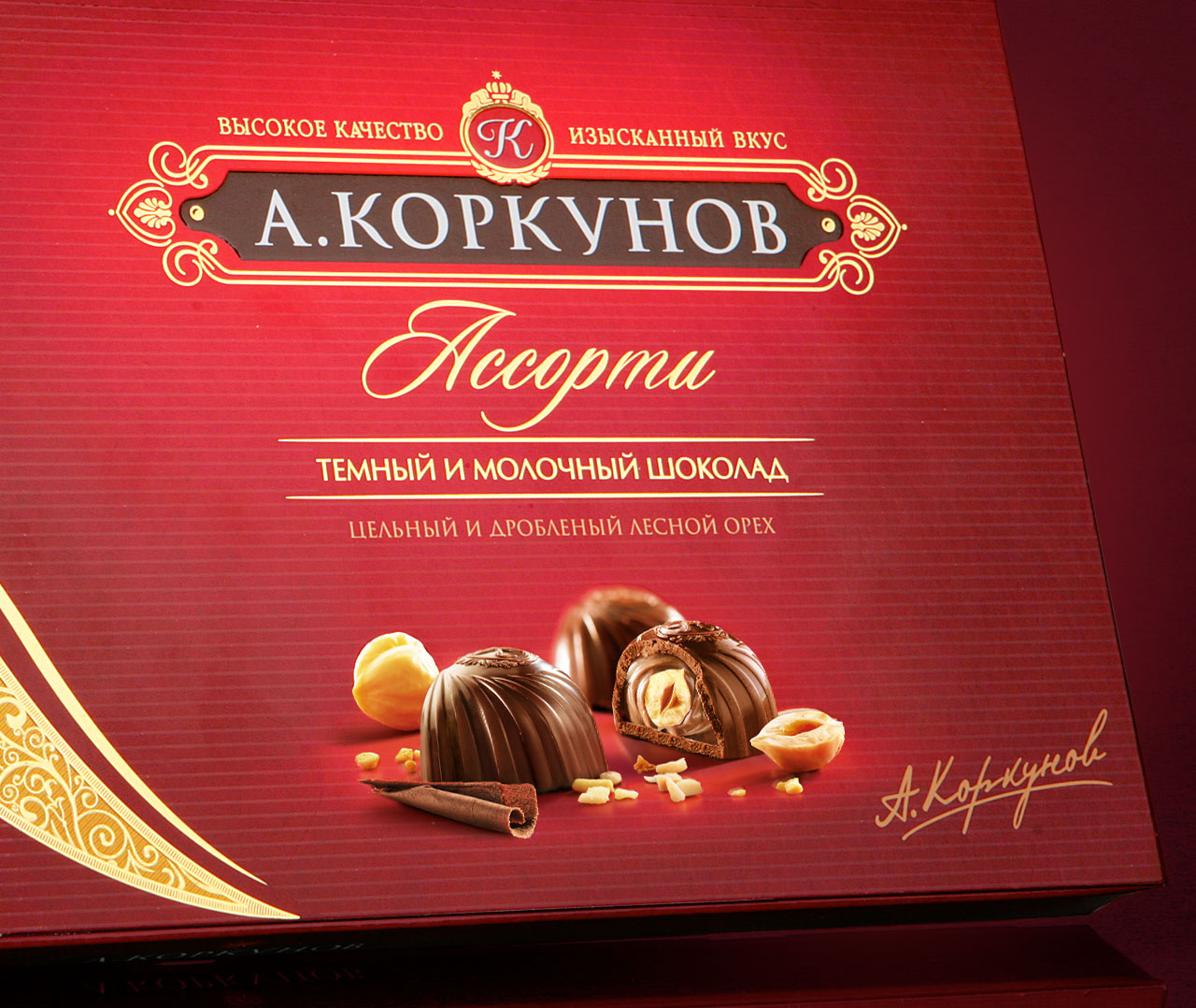 Красная коробка шоколадных конфет «Ассорти» А. Коркунова с изображениями целых и половинок шоколадных конфет с лесными орехами и шоколадным завитком, а также золотым и белым декоративным текстом на русском языке работы Солдиса.