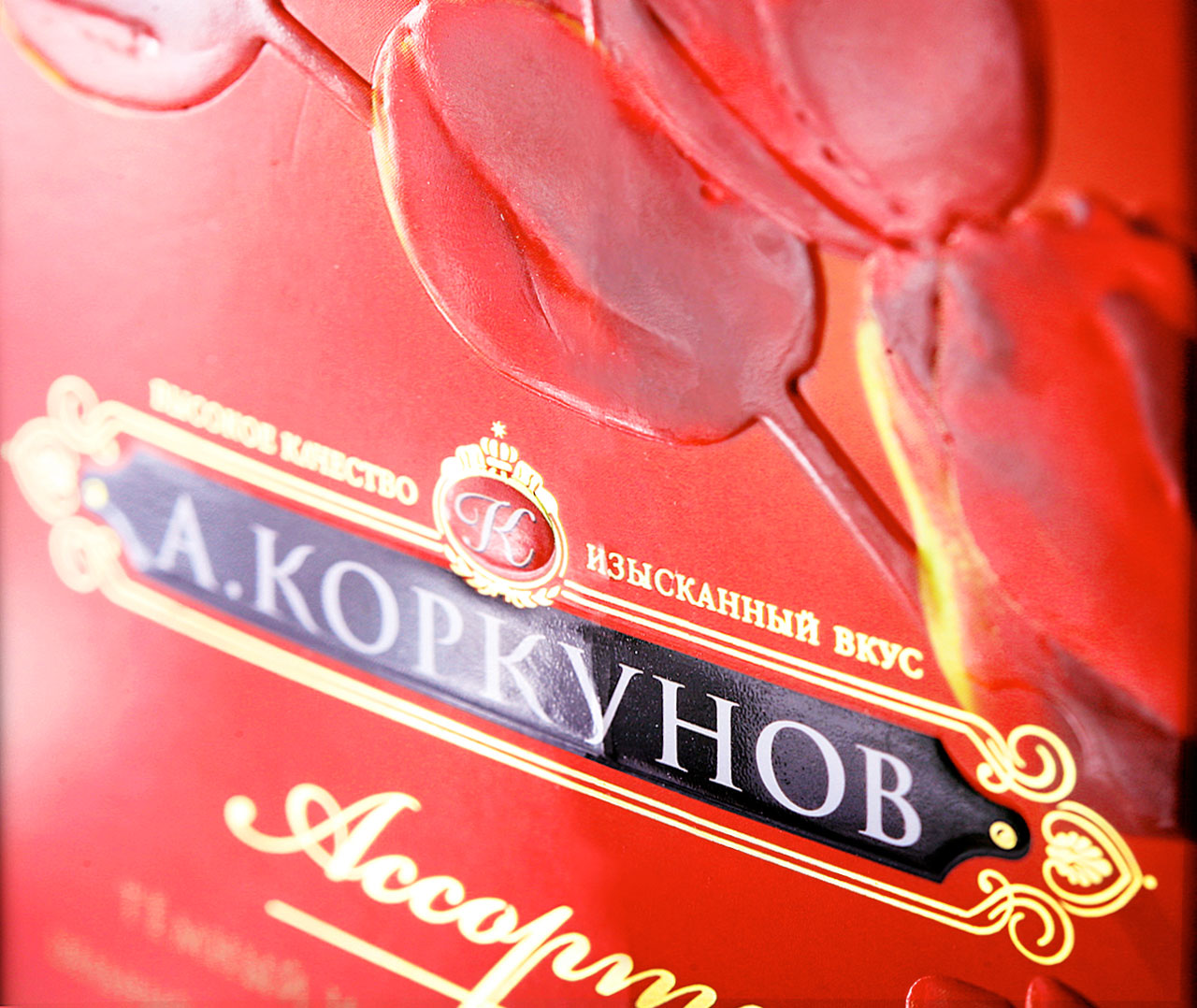 Крупный план красной коробки шоколадных конфет А. Коркунова с золотым и черным декоративным текстом на русском языке от Soldis, на заднем плане — рельефные изображения роз.