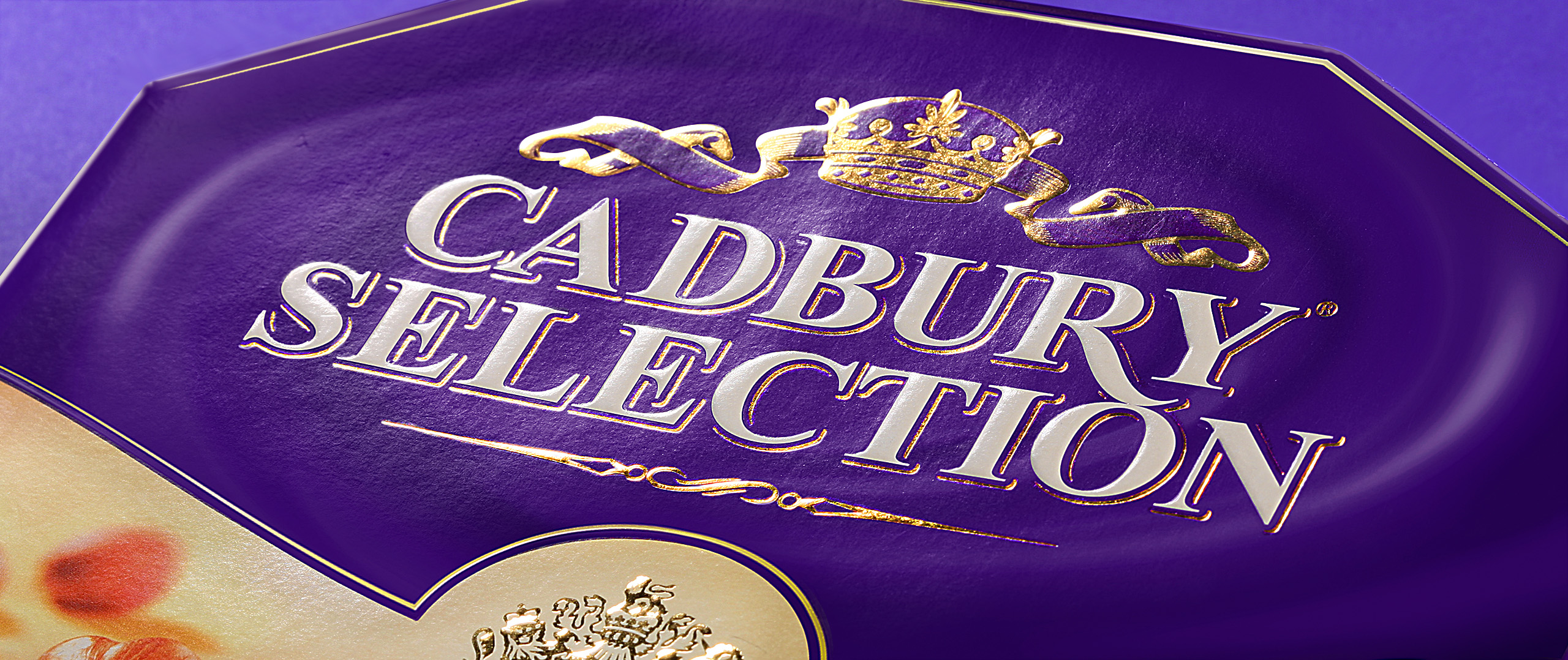 Крупный план фиолетовой коробки с надписью Cadbury Selection, написанной большими золотыми буквами, и декоративной короной над текстом. В нижнем углу виден край бежево-золотого дизайна Soldis.