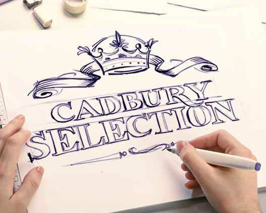 Человек рисует слова Cadbury Selection декоративными стилизованными буквами на белой бумаге с ручкой в руке. Дизайн Soldis включает ленту и корону над текстом, а также разбросанные по бумаге художественные принадлежности.